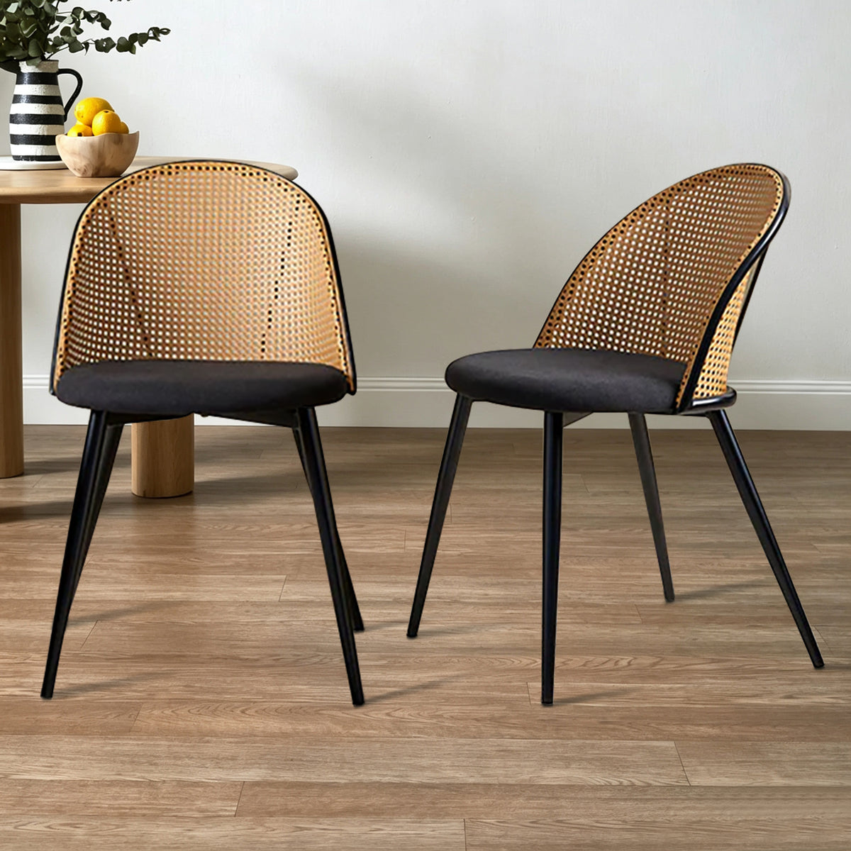 Two modern dining chairs with woven cane backrests and black upholstered seats, shown on a wood floor in a styled room setting.