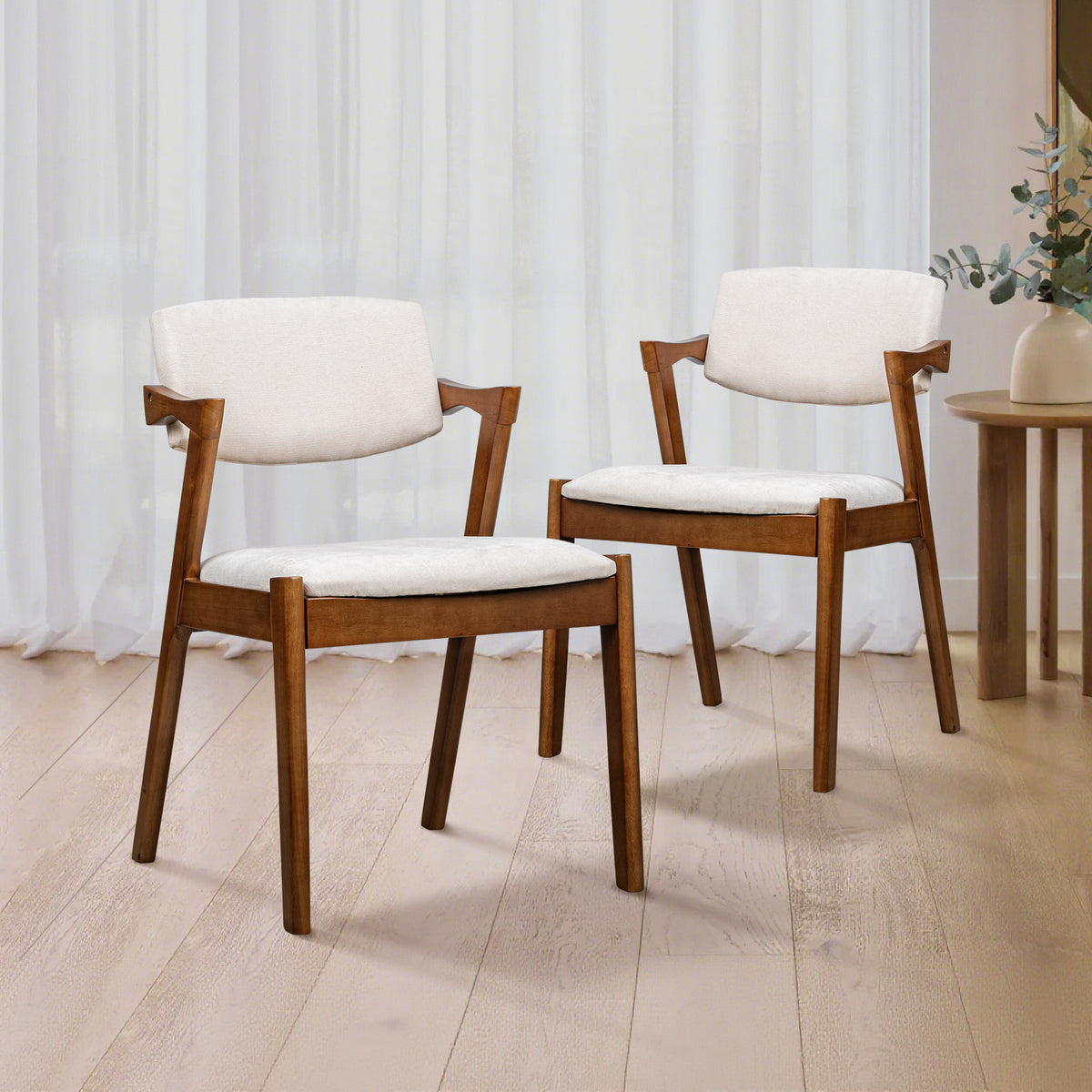 Two wooden dining chairs with white upholstered seats and curved armrests, shown in a bright room with light wood flooring and sheer white curtains.