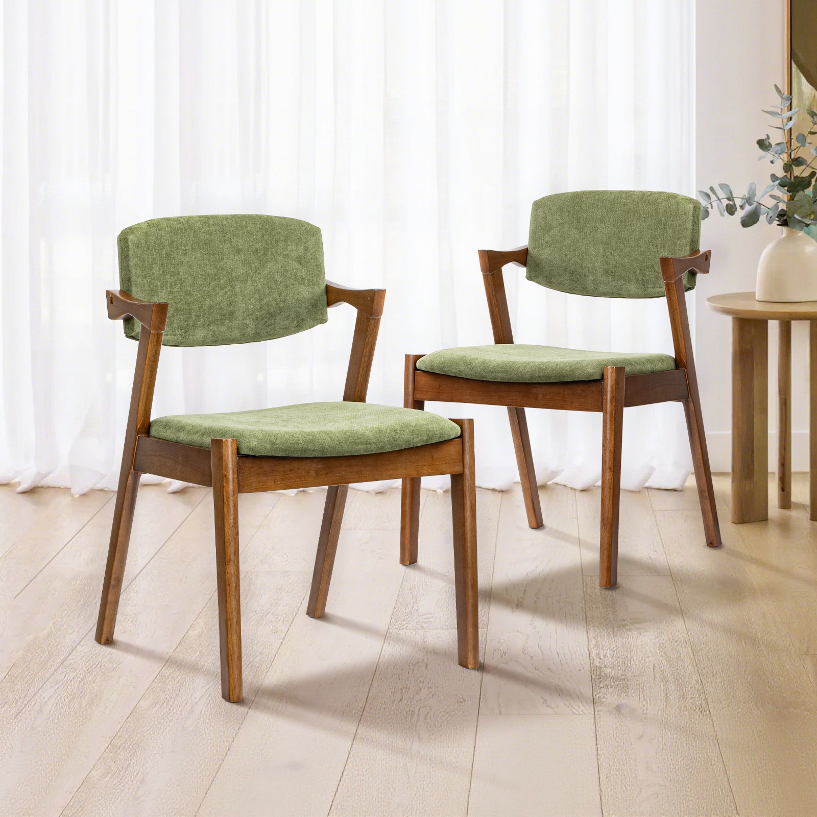 Two mid-century modern dining chairs with olive green velvet cushions and warm brown wooden frames, set on a light wood floor in front of white sheer curtains.