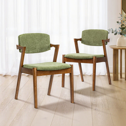 Two mid-century modern dining chairs with olive green velvet cushions and warm brown wooden frames, set on a light wood floor in front of white sheer curtains.