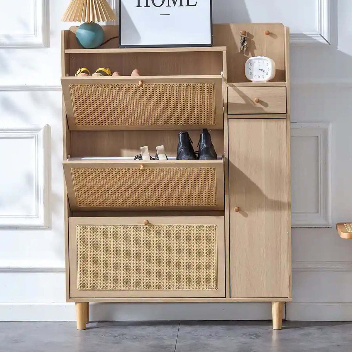 A light natural wood entryway cabinet with woven cane panels on the drawer fronts, shown in a styled room setting with shoes, a lamp, and wall decor.
