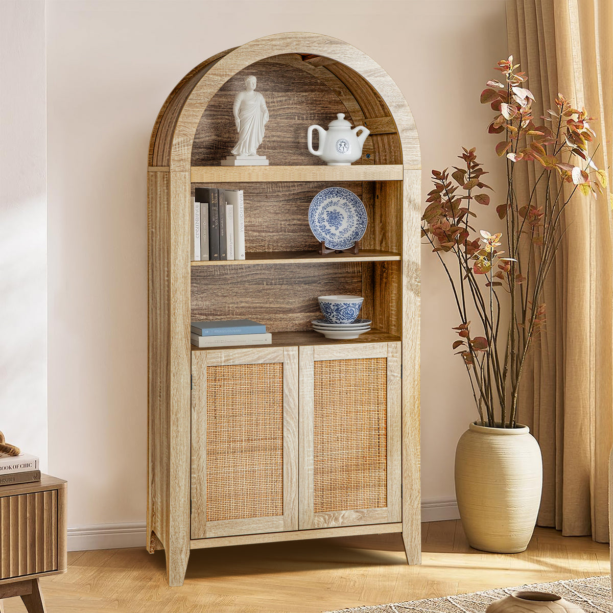 A tall rustic wooden cabinet with an arched top, open shelves, and two cane-embossed doors, styled in a warm beige room next to a large vase with dried branches.
