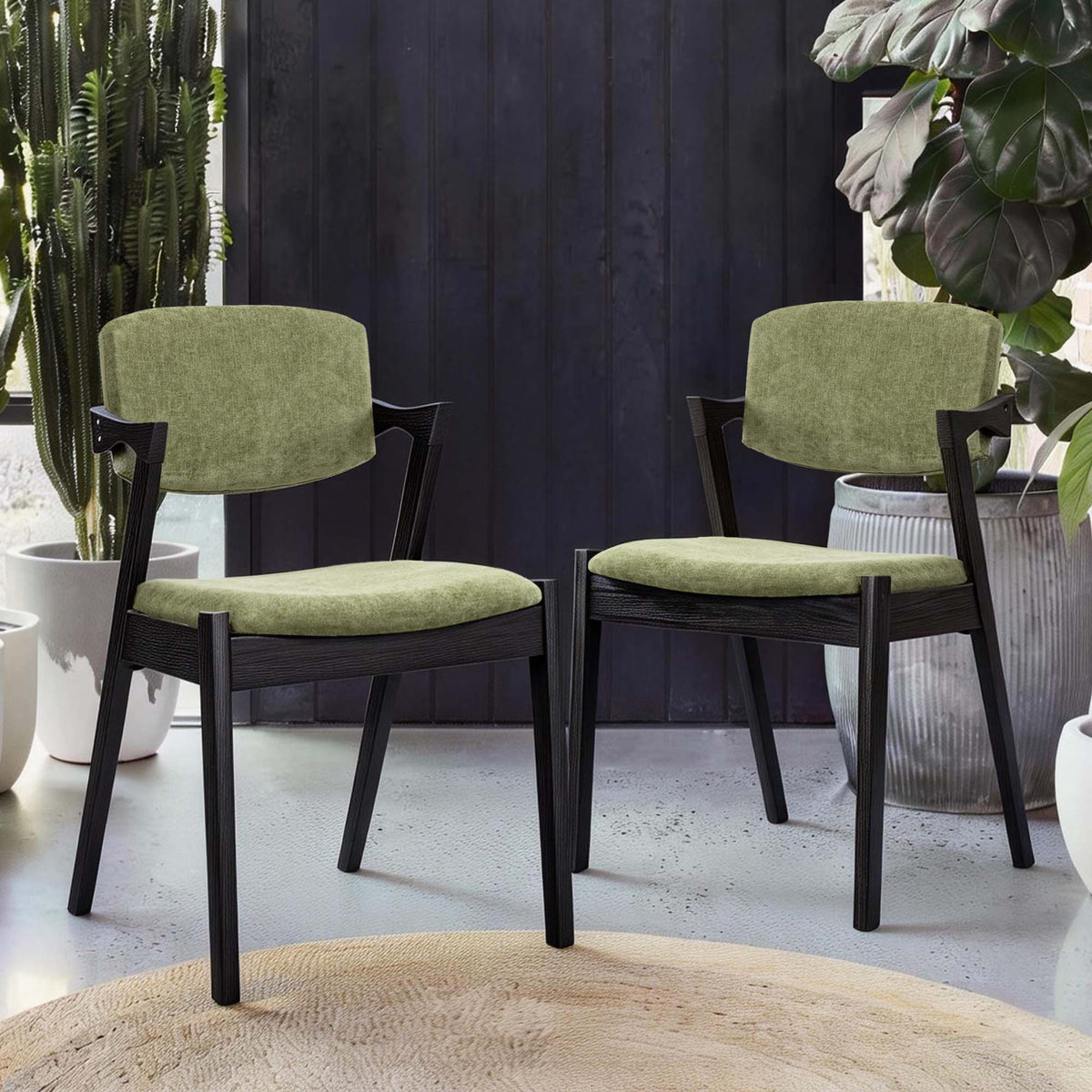 Two modern dining chairs with black wooden frames and light olive green velvet cushions, set in a styled interior with potted plants and a dark paneled wall.