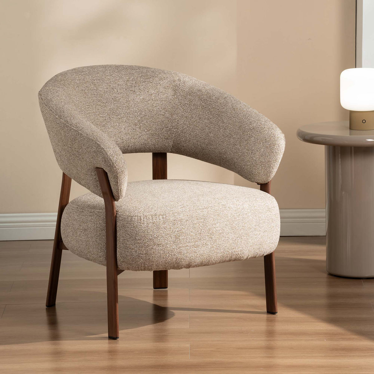A modern accent chair with a curved back and seat upholstered in textured beige fabric, supported by dark brown wooden legs, set in a room with a wooden floor and a side table.