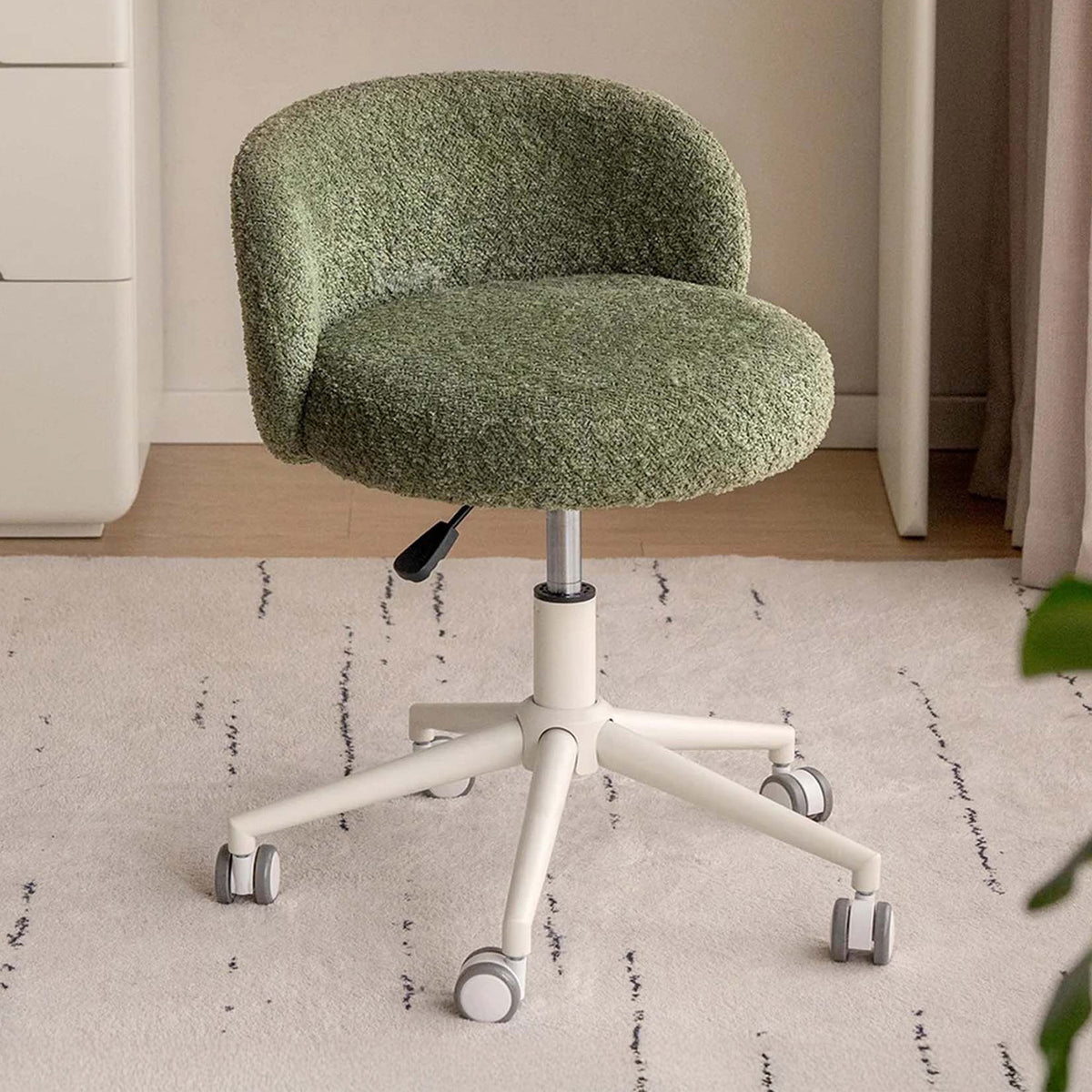 A small upholstered office chair with a textured sage green boucle-style seat and curved back, mounted on a white five-star wheeled base with a black lever, shown in a home office setting on a light rug.