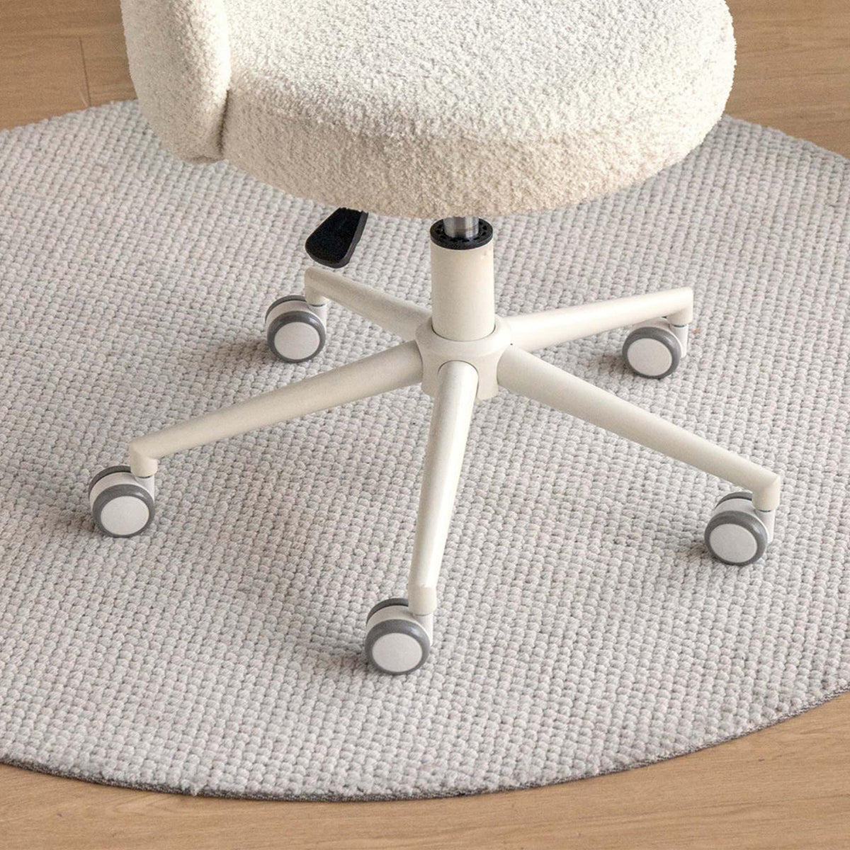 A close-up of a cream-colored, textured boucle office chair with a five-star wheeled base, positioned on a round, light gray woven rug over a light wood floor.