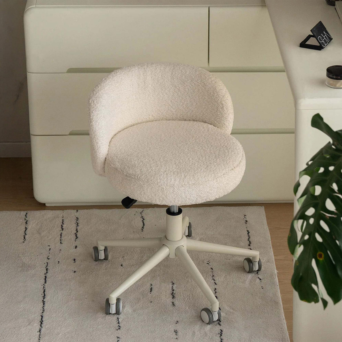 A cream-colored bouclé office chair with a five-star wheeled base, positioned on a light rug in front of a cream-colored dresser.