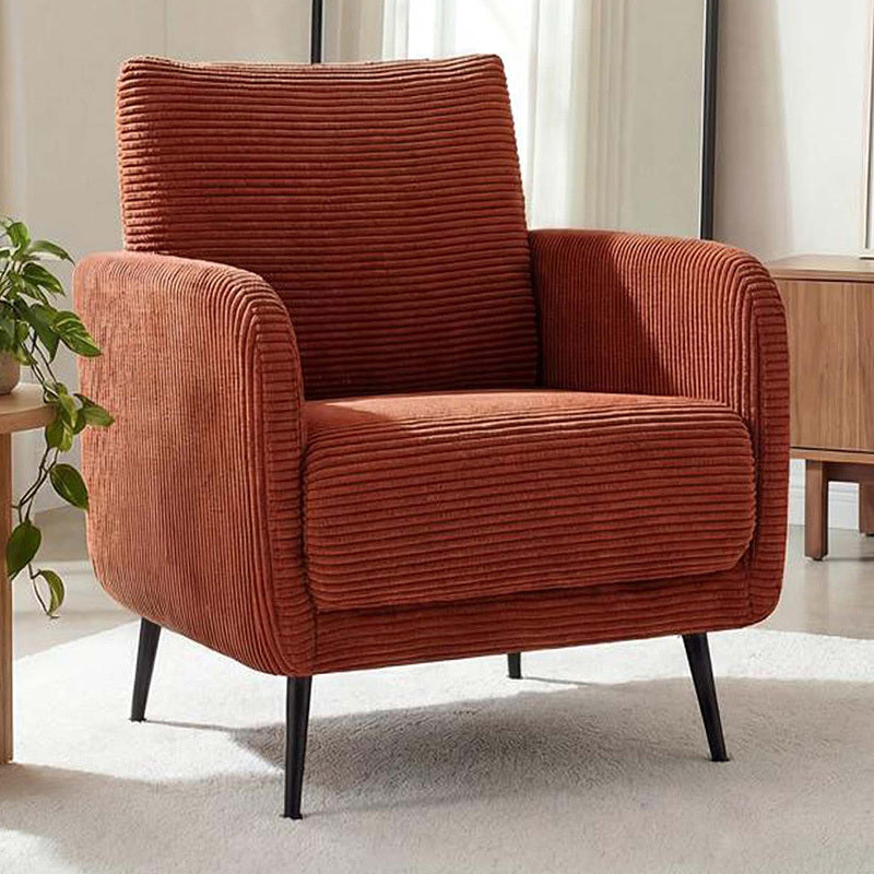 A modern corduroy armchair in a warm rust-orange color, styled in a bright living room setting with a light-colored rug, a wooden side table, and a potted plant.