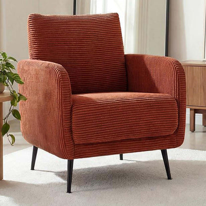 A modern corduroy armchair in a warm rust-orange color, styled in a bright living room setting with a light-colored rug, a wooden side table, and a potted plant.