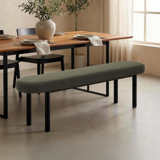 A long upholstered dining bench with a textured gray-green fabric seat and black metal legs, placed in front of a wooden dining table in a softly lit room.