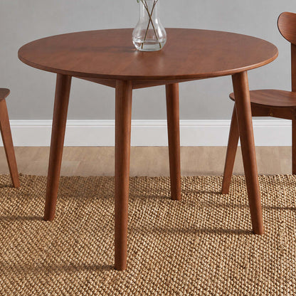A round wooden dining table with four tapered legs, shown in a styled room setting with a woven rug, chairs, and a glass vase on top.
