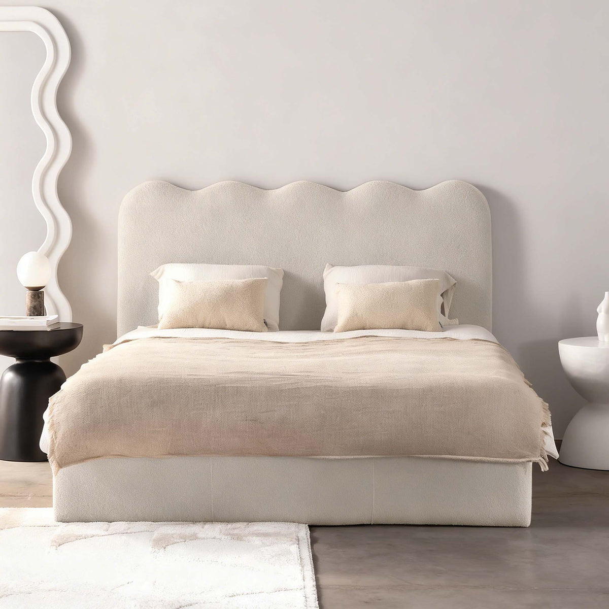 A modern upholstered bed with a scalloped headboard, dressed in neutral beige linens and two matching pillows, set in a minimalist bedroom with a sculptural white side mirror and dark side table.