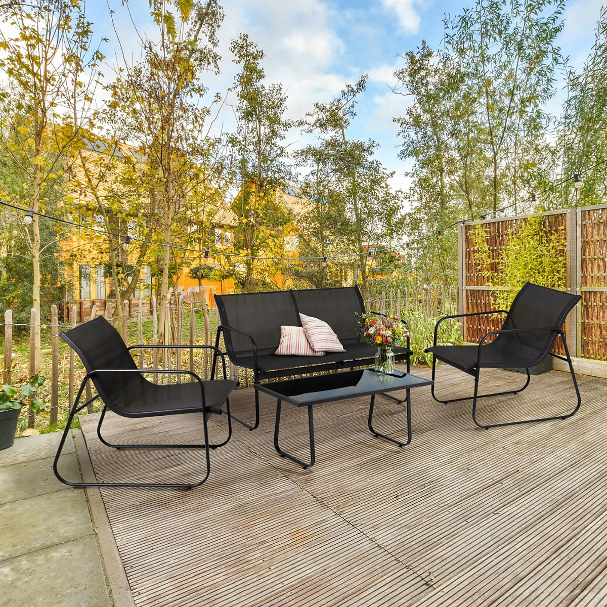 A 4-piece black outdoor patio furniture set including a 3-seat sofa, two armchairs, and a coffee table, arranged on a wooden deck in a backyard setting with trees and a fence.