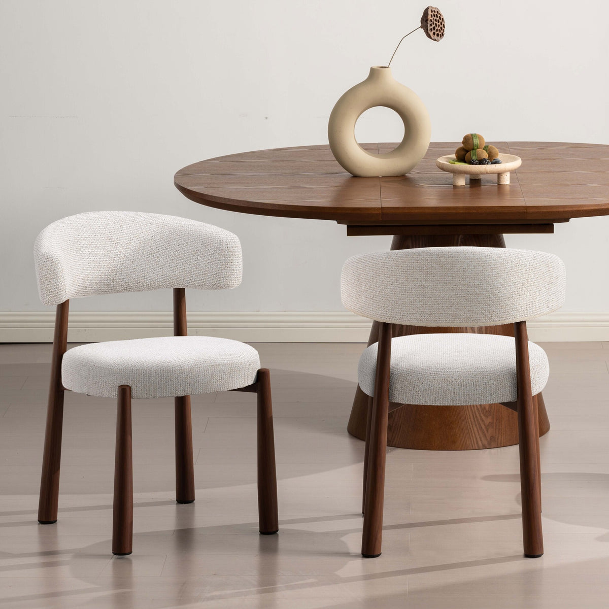 Two upholstered dining chairs with cream boucle-style seats and backs and dark wood legs, shown beside a round wooden dining table in a clean interior setting.