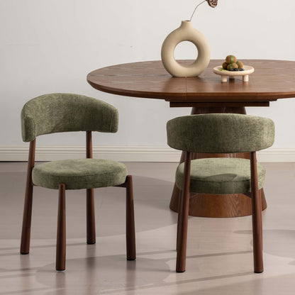 Two upholstered dining chairs with olive green fabric and dark wood legs, shown beside a round wooden dining table in a minimalist room setting.