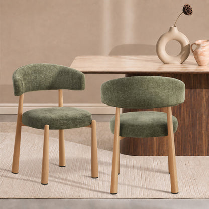 Two upholstered dining chairs with sage green fabric seats and curved backs, supported by light natural wood legs, shown in a styled dining room setting next to a wooden table.