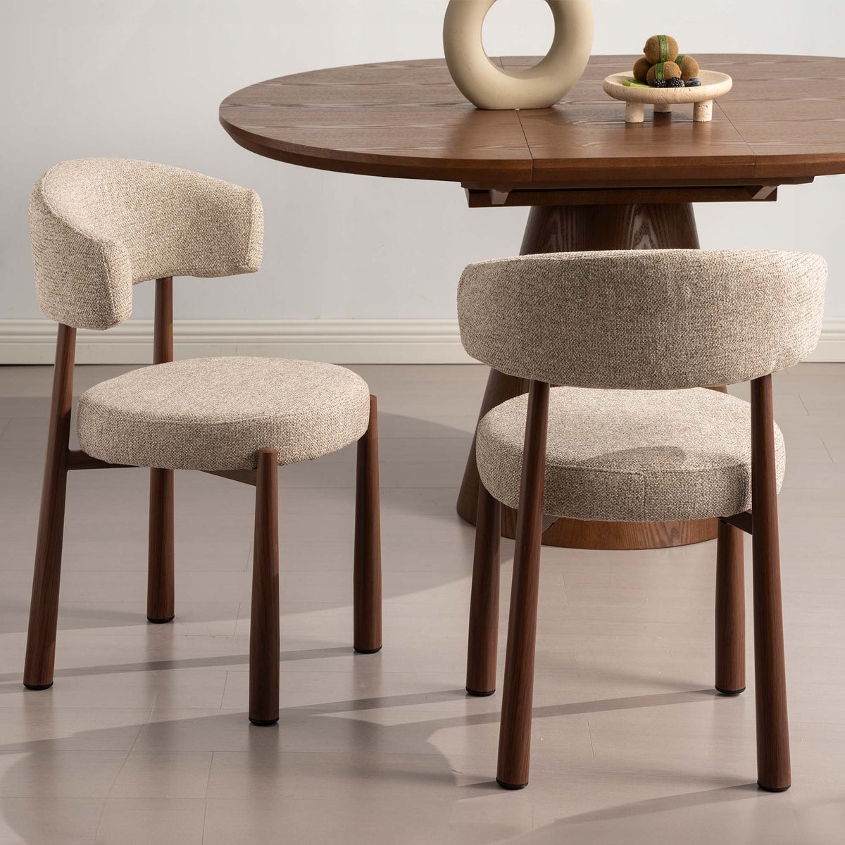 Two upholstered dining chairs with curved backs and round seats in a textured beige bouclé fabric, paired with a round wooden dining table in a bright, modern room.
