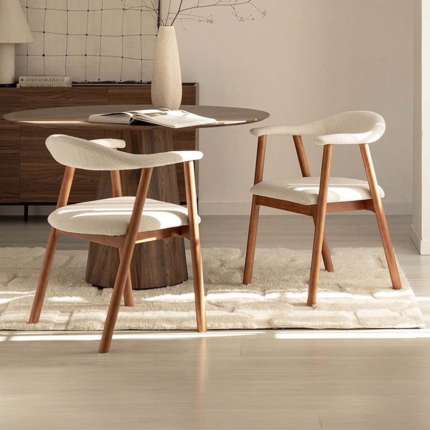 Two modern dining chairs with cream upholstered seats and curved wooden legs, arranged around a round wood dining table in a bright, minimalist dining room.