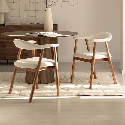 Two modern dining chairs with cream upholstered seats and curved wooden legs, arranged around a round wood dining table in a bright, minimalist dining room.