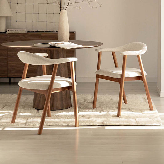 Two modern dining chairs with cream upholstered seats and curved wooden legs, arranged around a round wood dining table in a bright, minimalist dining room.