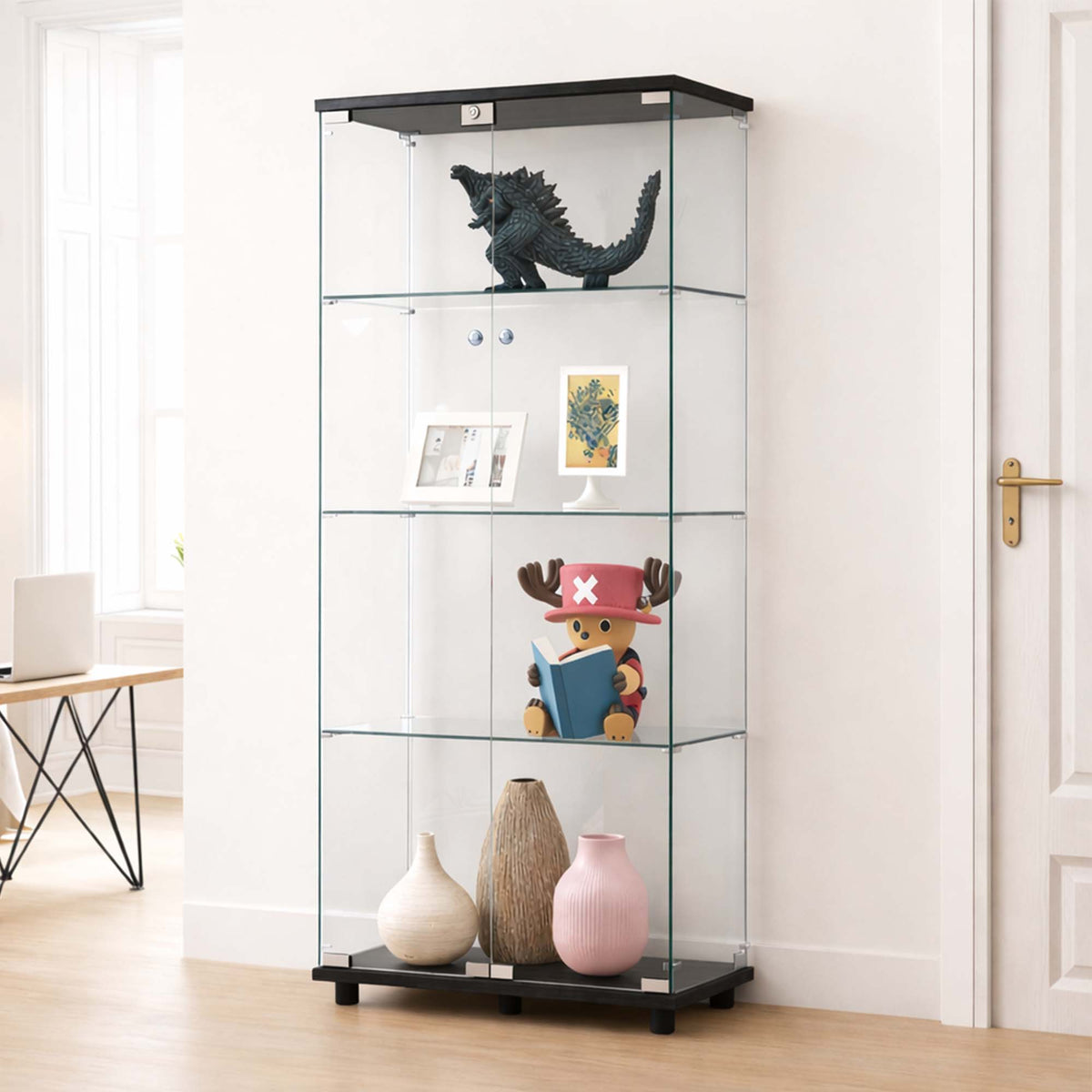 A tall glass display cabinet with black top and bottom frames, shown in a bright room with shelves holding a dinosaur figurine, framed art, a small plush toy, and three vases.